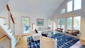 A spacious living room with a large sofa, a glass coffee table, and a blue patterned rug. There is a staircase on the left and large windows on the right. - Hot Tub, Fireplace & Pet Friendly! Come Visit "Lake It or Knot"! (Huddleston)