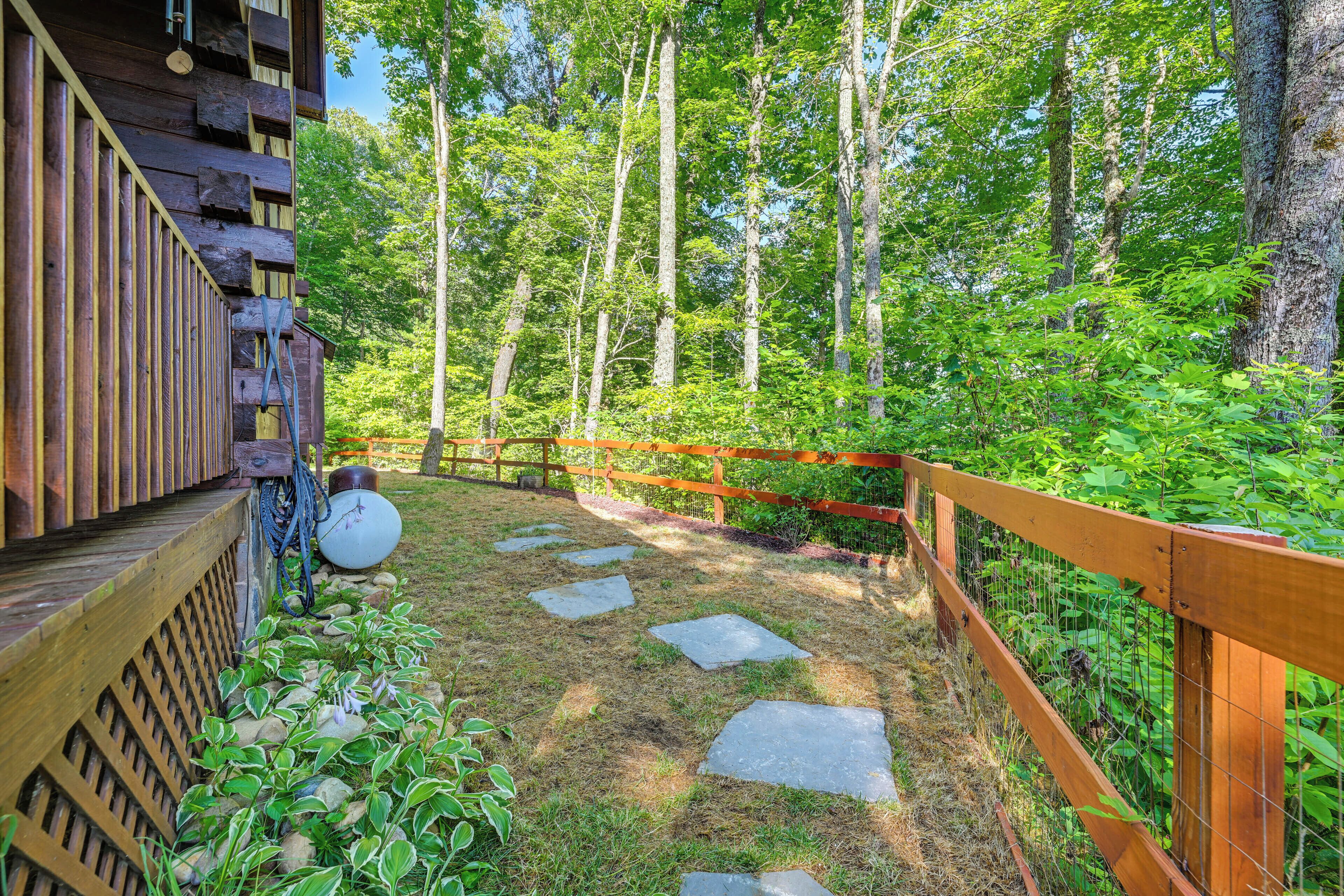 Una terraza de madera con un sendero de piedra que conduce a un área cercada, rodeada de abundante vegetación y árboles altos.