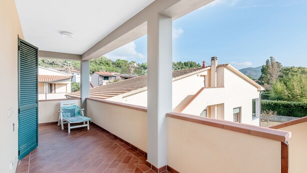 A balcony with a tiled floor, a white chair, and a view of neighboring buildings and greenery. - Just 500 m from the sandy beach - Primula 2 (Marina di Campo)