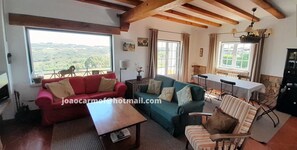 A cozy living room with a red sofa, green sofa, and a striped armchair. There is a wooden coffee table, a dining table with chairs, and a black cabinet. Large windows offer a view of the countryside.