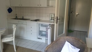 Una cocina compacta con armarios blancos, lavavajillas integrado y cafetera. En primer plano, una silla de mimbre con un cojín.