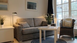 Una sala de estar con un sofá gris, una mesa de centro blanca, un sillón de mimbre y un ventanal con cortinas.