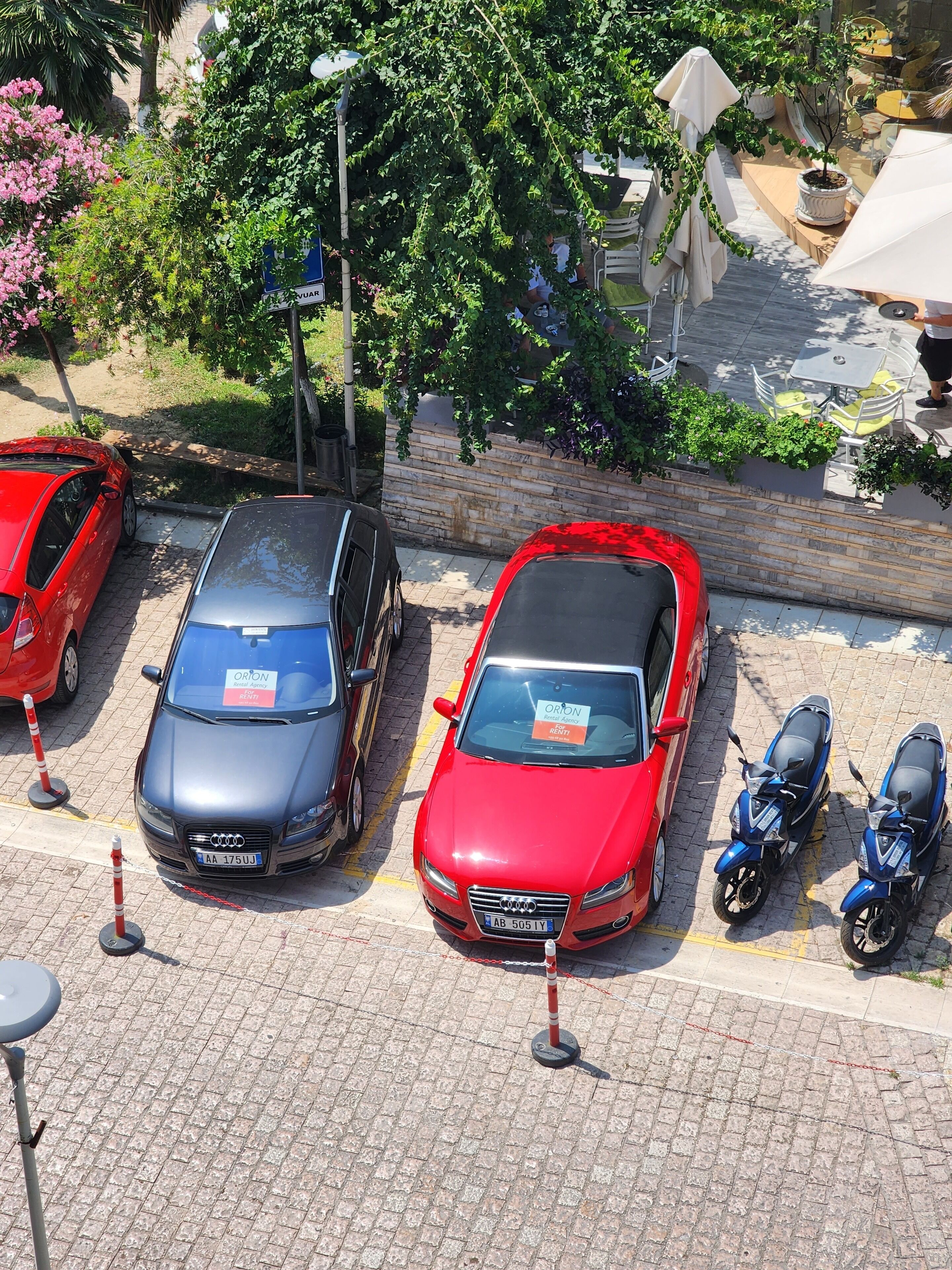 Ein Parkplatz mit Autos, Rollern, einer Steinmauer und einigen Grünpflanzen.