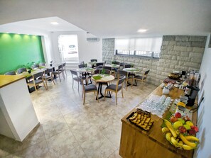 Uma sala de jantar moderna com várias mesas e cadeiras, um balcão de cozinha com frutas e bebidas, e uma parede verde.