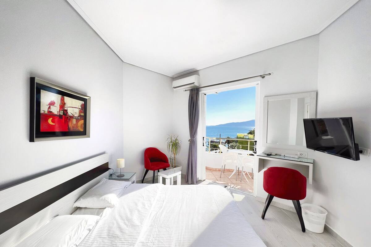 Habitación de hotel con cama, escritorio con computadora, televisor, una silla roja, una mesita de noche con lámpara y ventana con vista al mar y a las montañas.