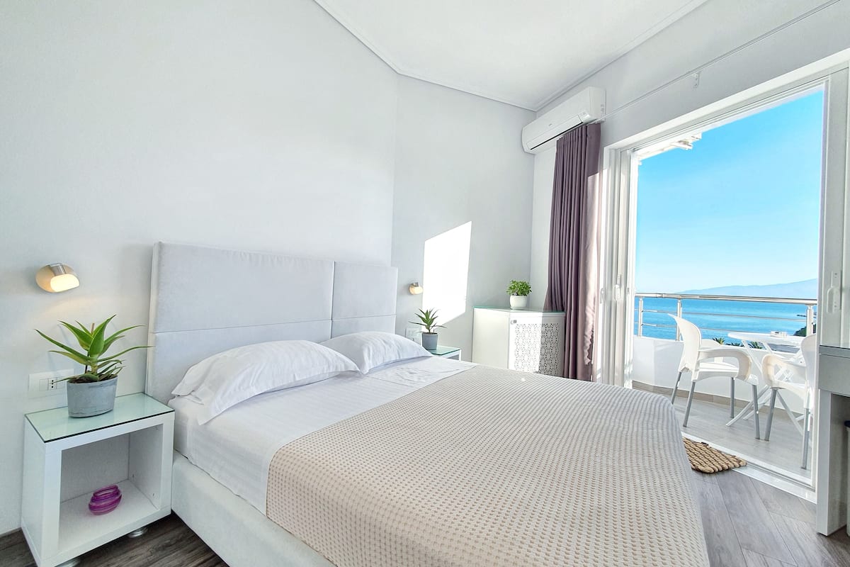 Un dormitorio moderno con una cama grande, una mesita de noche con lámpara, una planta en maceta y vistas al mar desde un balcón.