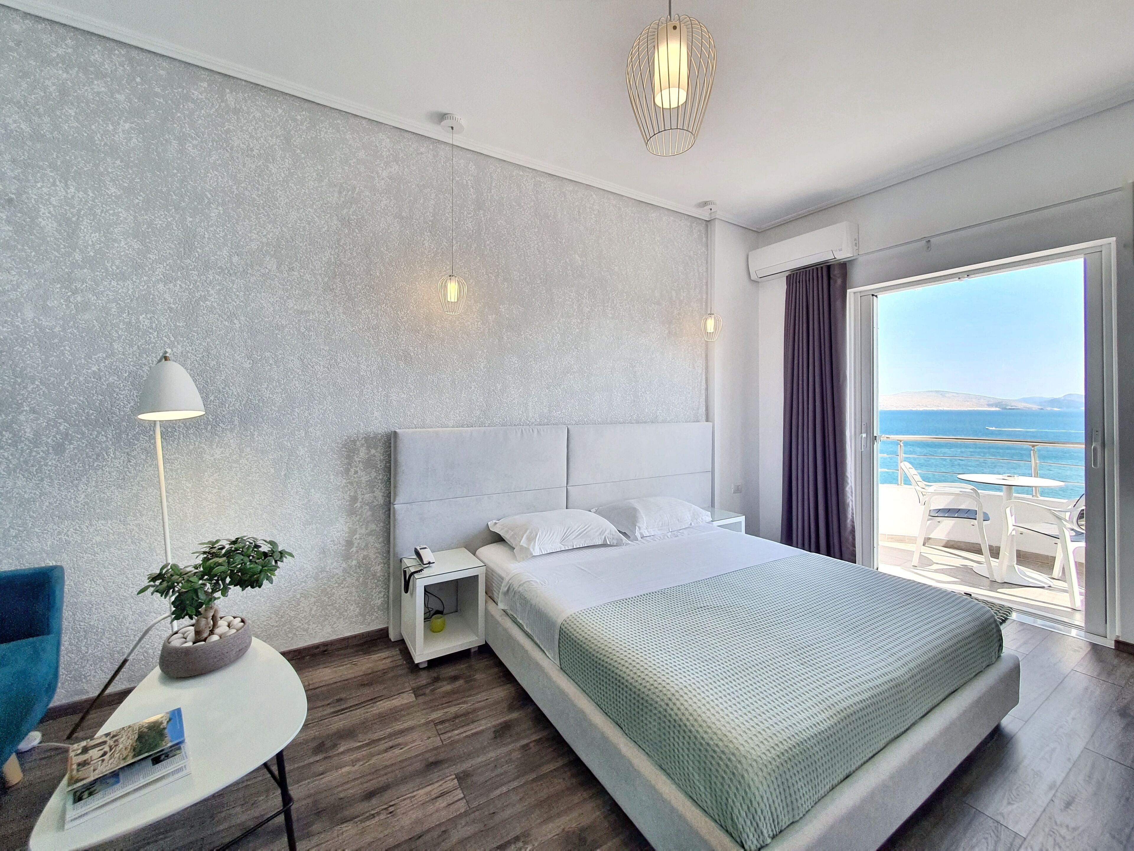 A modern hotel room with a large bed, a small bedside table with a lamp, a potted plant, and a view of the sea through a large window.
