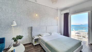 A modern hotel room with a large bed, a small bedside table with a lamp, a potted plant, and a view of the sea through a large window.