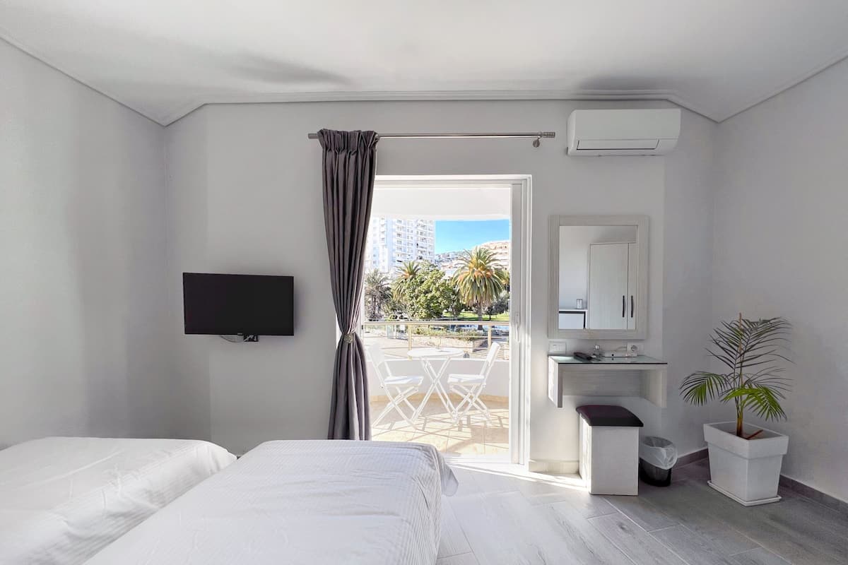 Habitación de hotel con dos camas, televisión, baño con lavabo y espejo, y balcón con vistas a edificios y palmeras.