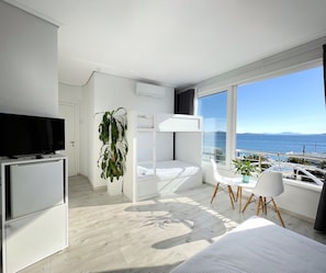 Quarto de hotel moderno com beliche, televisão sobre um móvel, planta em vaso e vista para o mar.