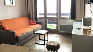 A modern living room with a grey sofa, a small table, and a stool. There is a window with curtains, a flat-screen TV, and a lamp on a desk.