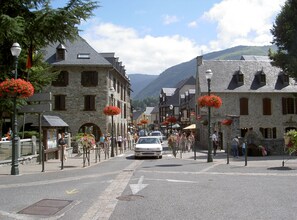 Une rue historique bordée de bâtiments en pierre, avec une voiture blanche et des jardinières rouges accrochées aux lampadaires.