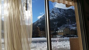 Vista nevada de una montaña a través de una ventana con cortinas, mostrando un edificio con varias ventanas y una calle con coches estacionados.