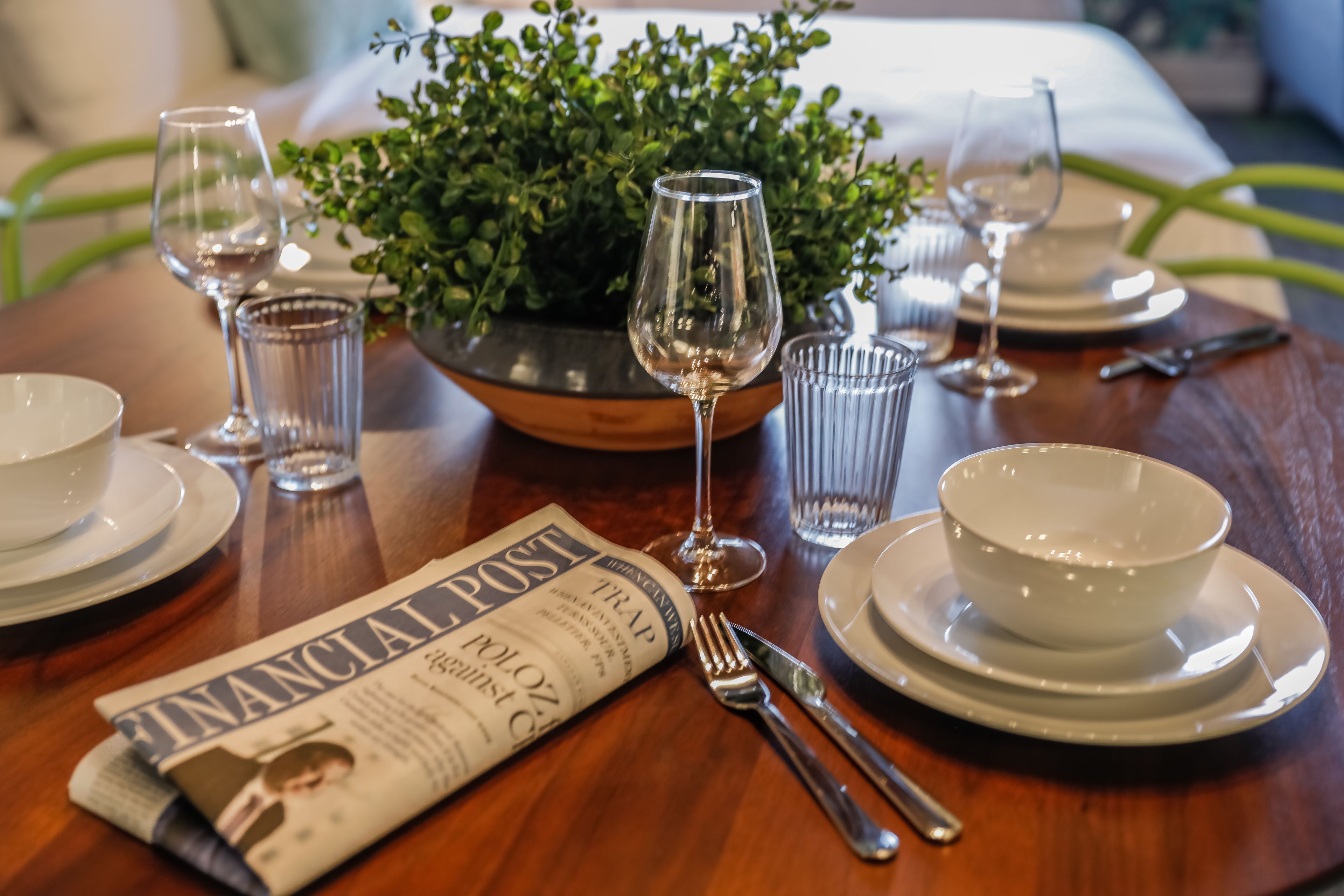 A dining table set with white plates, bowls, cutlery, and wine glasses. A newspaper is placed on the table.