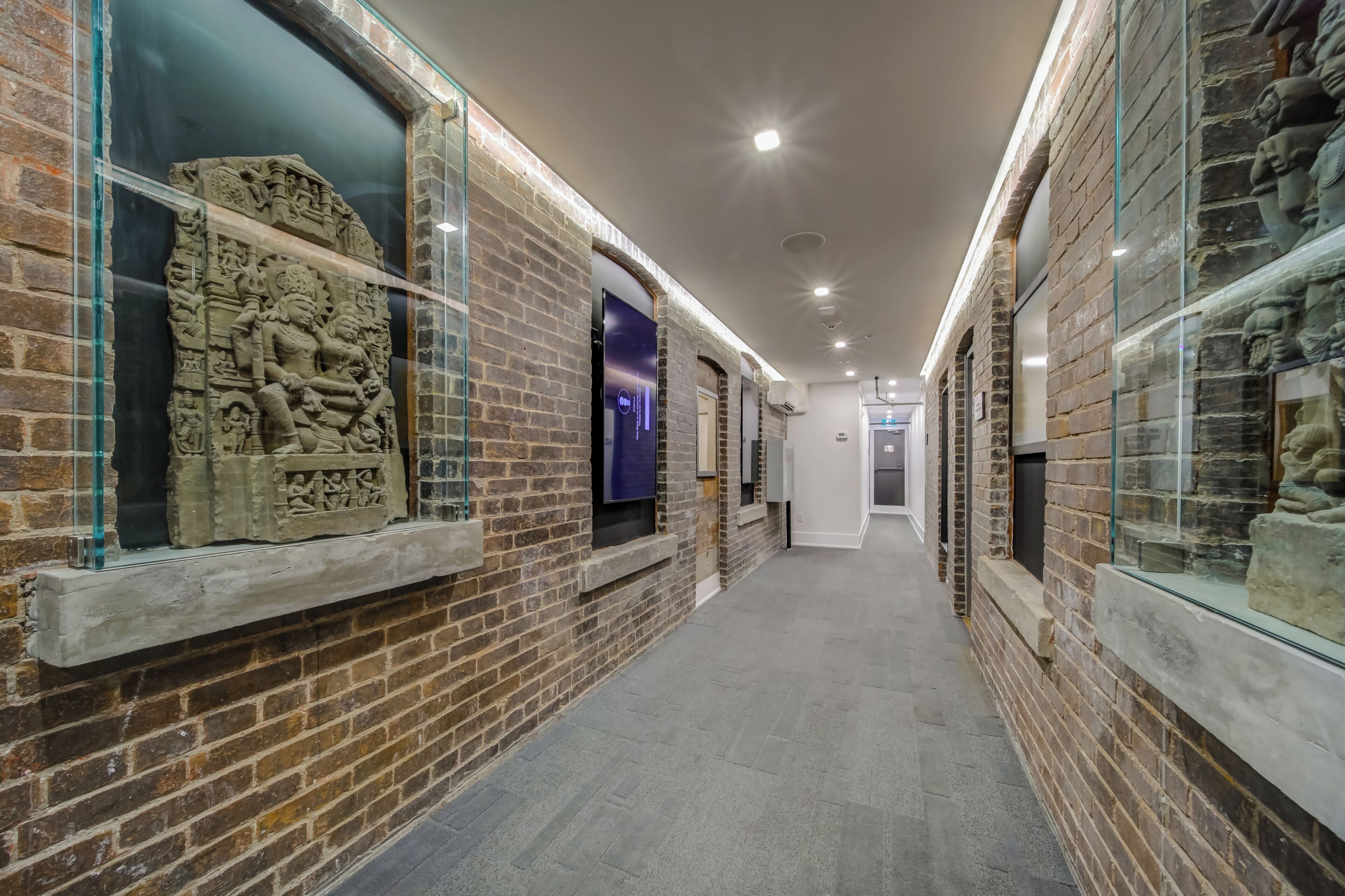 A corridor with brick walls, glass display cases with stone carvings, and a modern ceiling with recessed lighting.