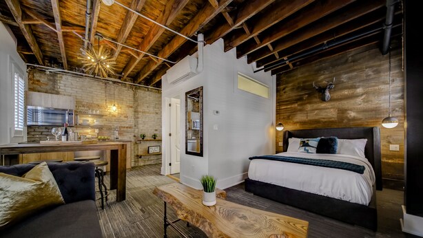 A modern, rustic room with a wooden ceiling, a large bed with a dark headboard, a wooden bench, and a small table with a plant. - The Westlake (Toronto)