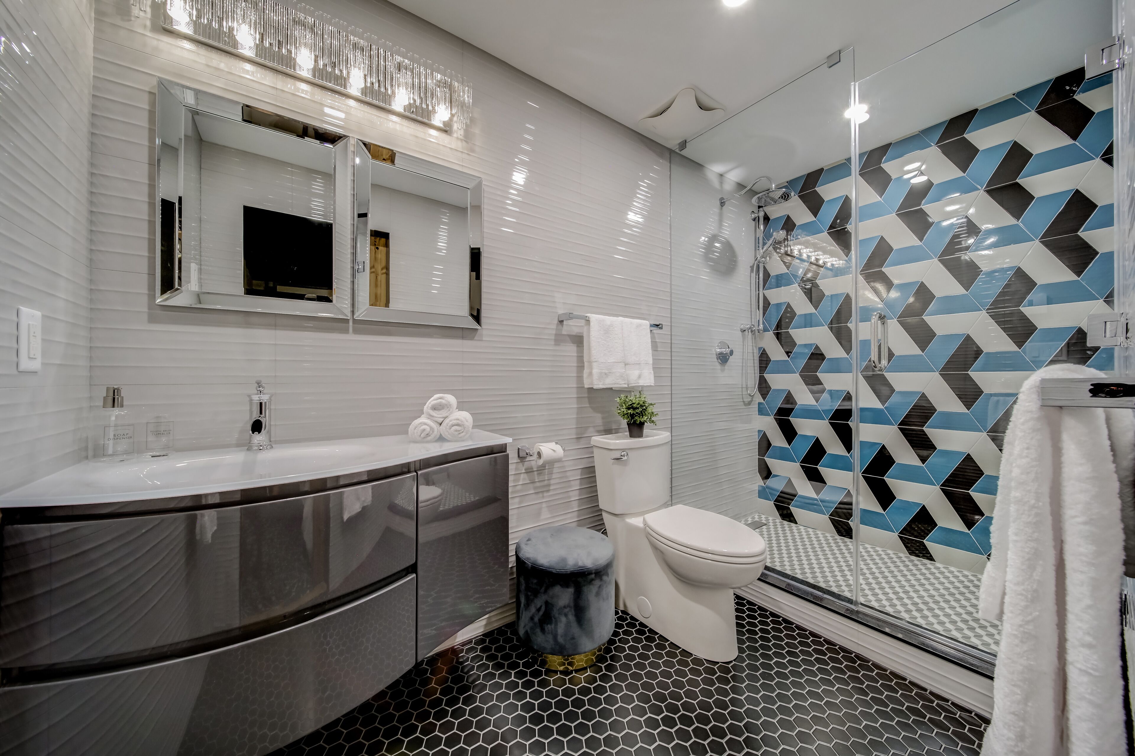 A modern bathroom with a large mirror, a white sink, a toilet, and a shower with a geometric tile pattern.