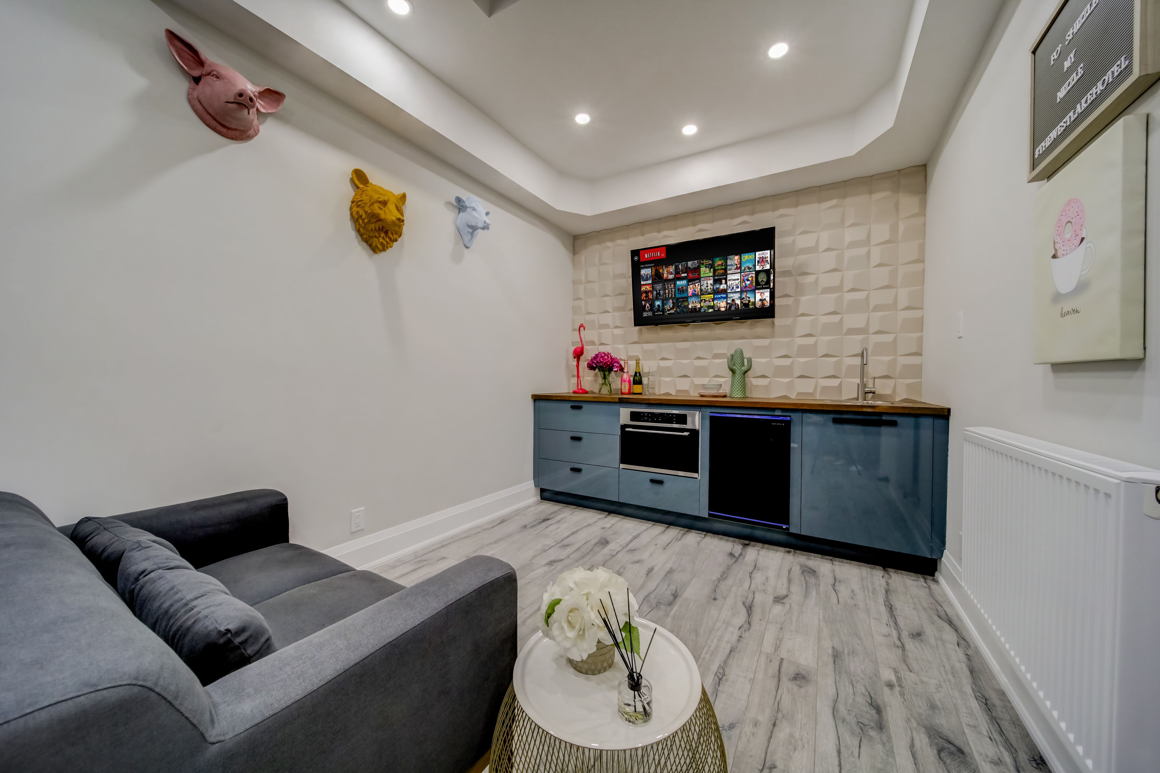 Un salon moderne avec un canapé gris, une petite table ronde ornée d’un vase, et un coin cuisine équipé d’une télévision et d’un plan de travail en bois.
