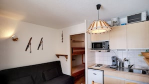A compact living space with a sofa, a dining table with a patterned tablecloth, a kitchen area with a microwave, a refrigerator, and a hanging light fixture.