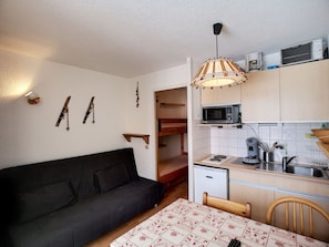 Um espaço compacto com sofá, mesa de jantar com toalha de mesa estampada, área de cozinha com micro-ondas, geladeira e luminária pendente.