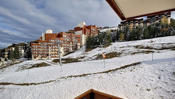 A snow-covered landscape with modern multi-story buildings, likely a hotel or resort, featuring balconies and a ski slope in the background.