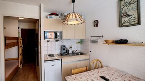 A compact kitchen with a refrigerator, microwave, and coffee maker. There is a dining table with a patterned tablecloth and a remote control on it. A wooden chair is placed at the table.