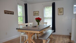 Un coin repas avec une table en bois, des chaises blanches et un banc. Il y a deux fenêtres équipées de stores et un vase avec des fleurs rouges sur la table.