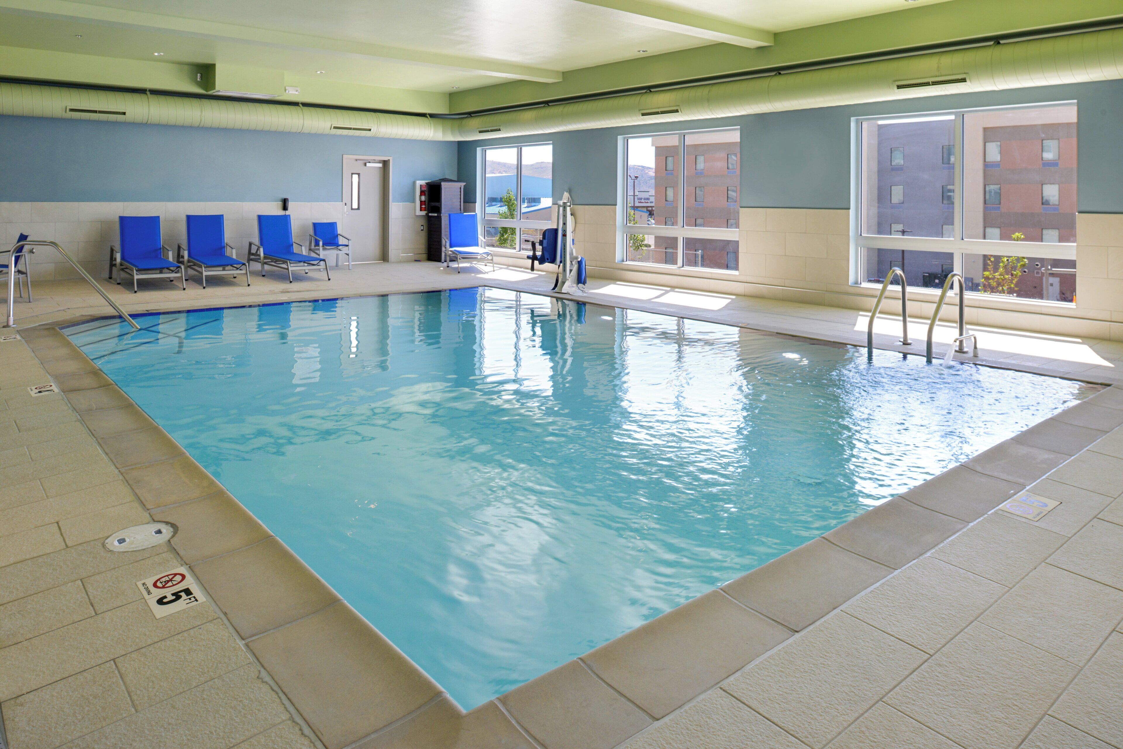 An indoor swimming pool with clear blue water, surrounded by blue chairs and a window with a view of buildings.
