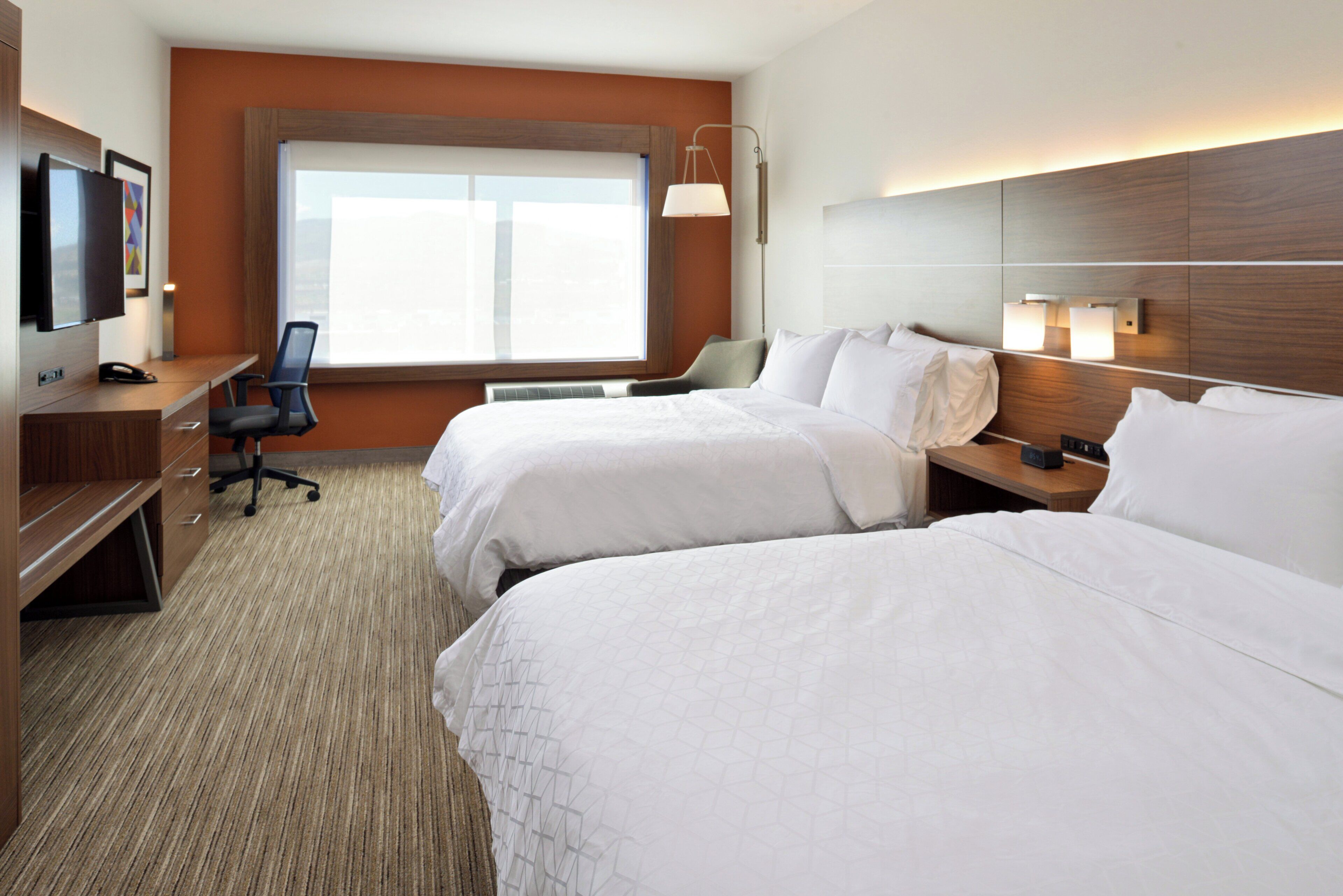 A hotel room with two beds, a desk, a chair, a TV, and a window.
