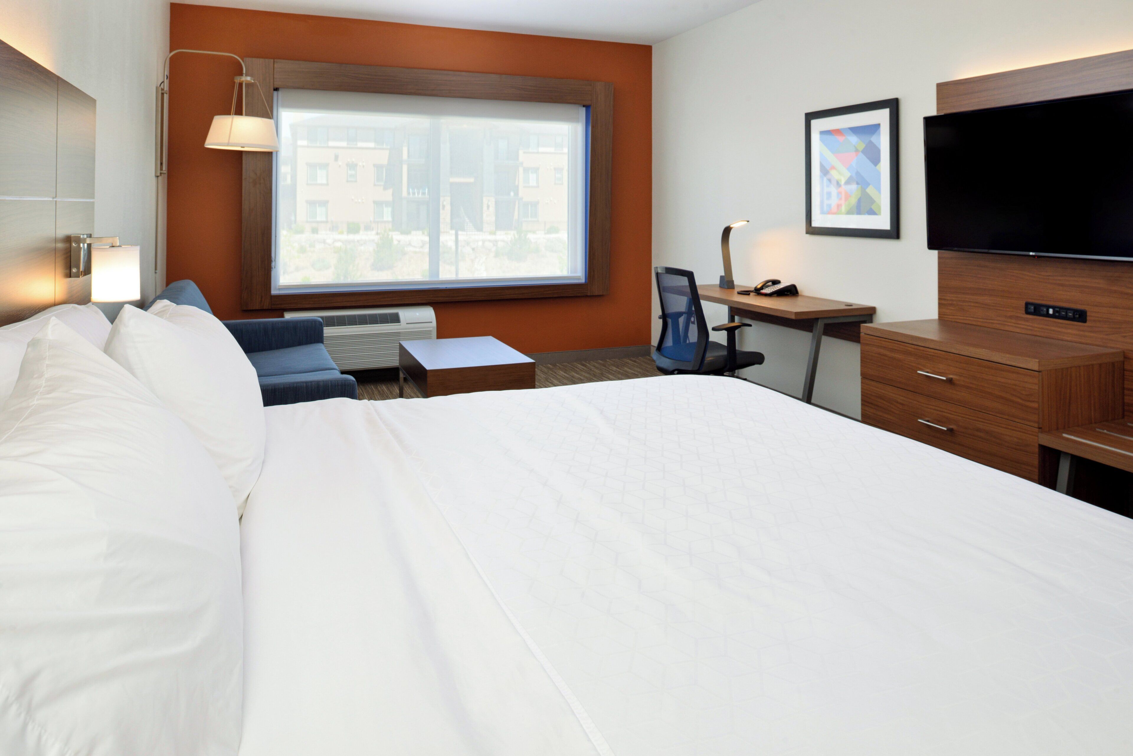 A hotel room with a large bed, a desk, a chair, a flat-screen TV, and a window with a city view.