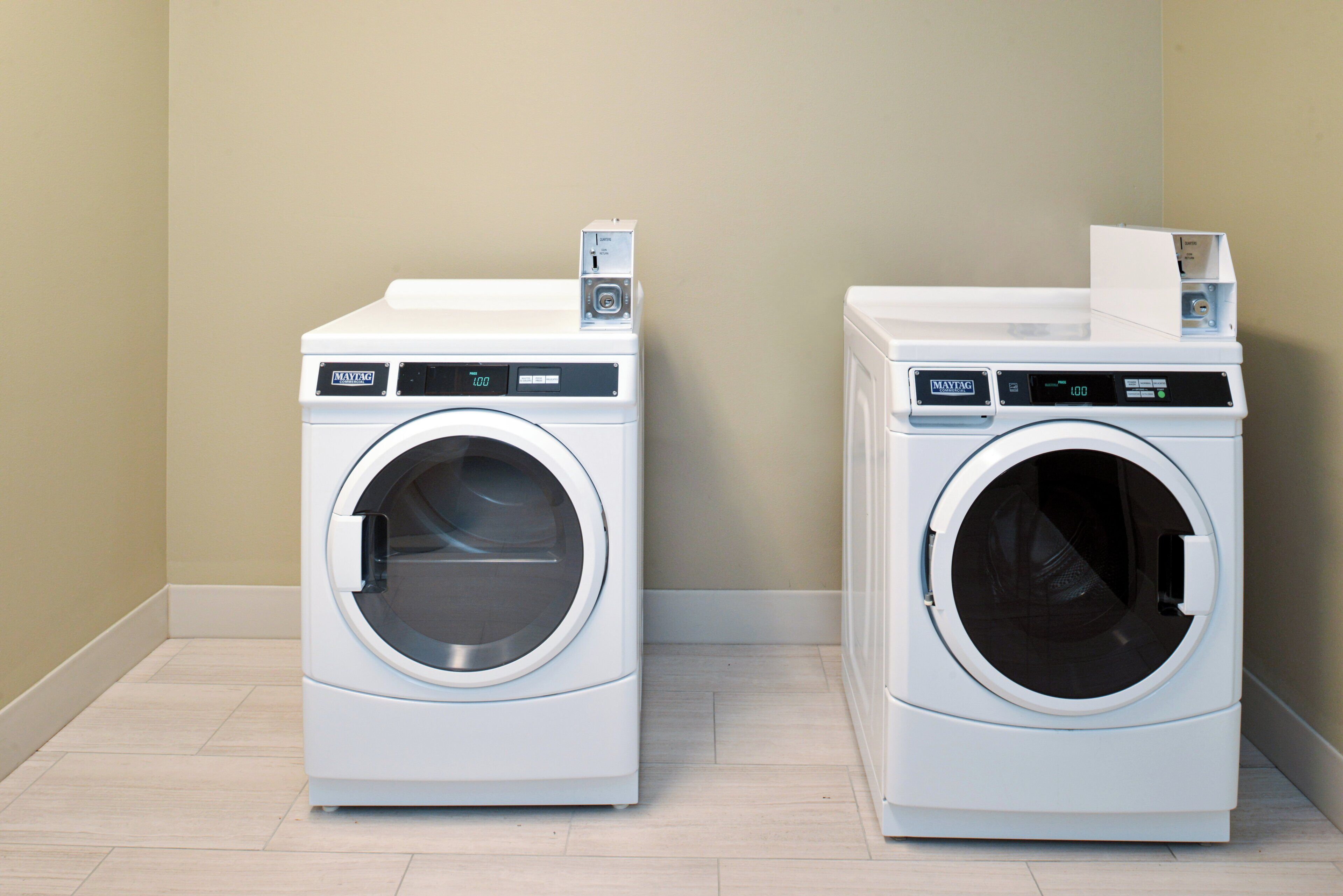 Two white Maytag commercial washing machines in a corner with a beige wall and tiled floor.
