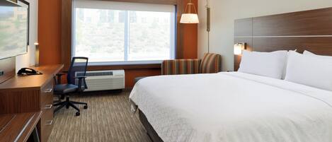 A hotel room with a large bed, a desk with a chair, a television, and a window with a view of trees.