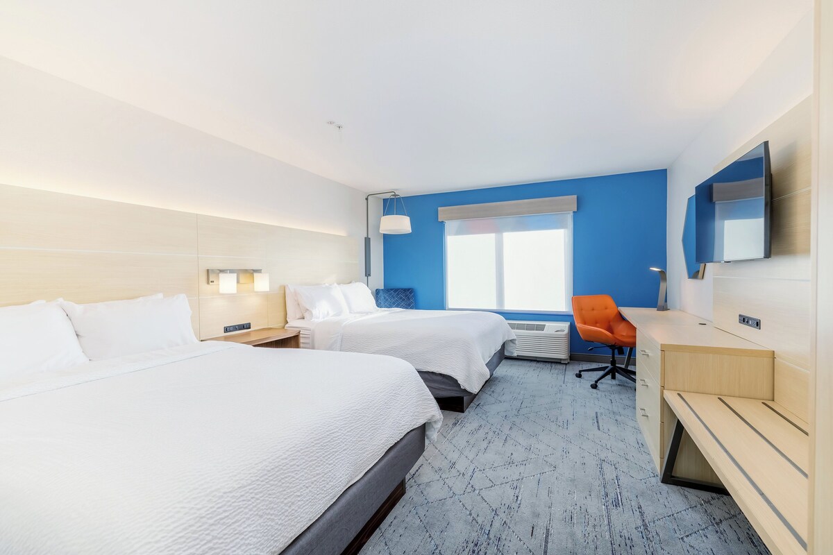 A hotel room with two beds, a desk, an orange chair, a flat-screen TV, and a large window.