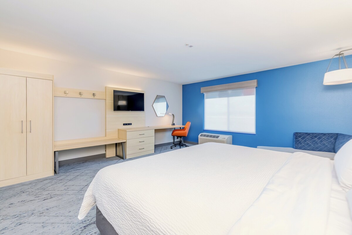 A bedroom with a bed, a desk, a chair, a TV, and a window.