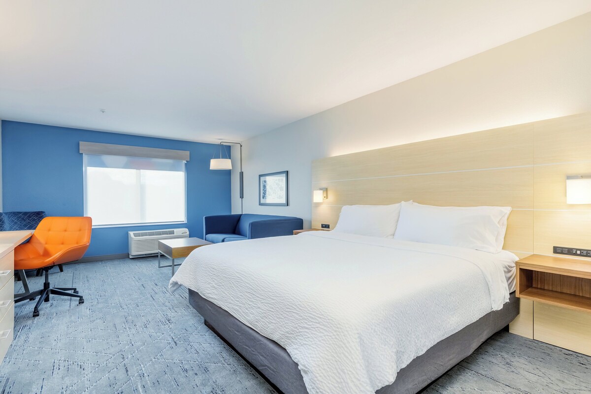 A hotel room with a large bed, a desk with a chair, a window with blinds, and a wall-mounted light.