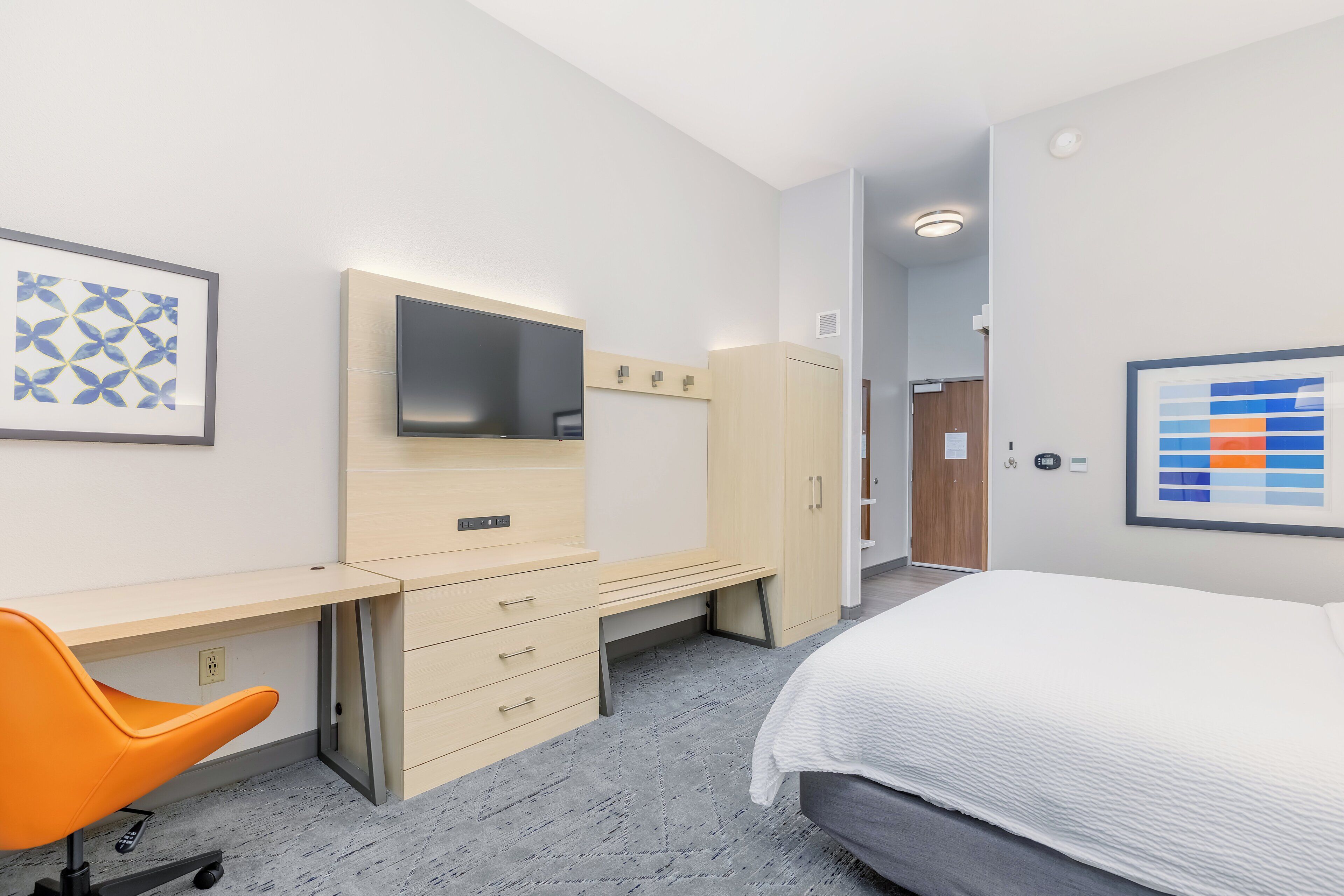 A hotel room with a bed, a desk with a chair, a wardrobe, a TV, and artwork on the walls.