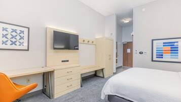 A hotel room with a bed, a desk with a chair, a wardrobe, a TV, and artwork on the walls.