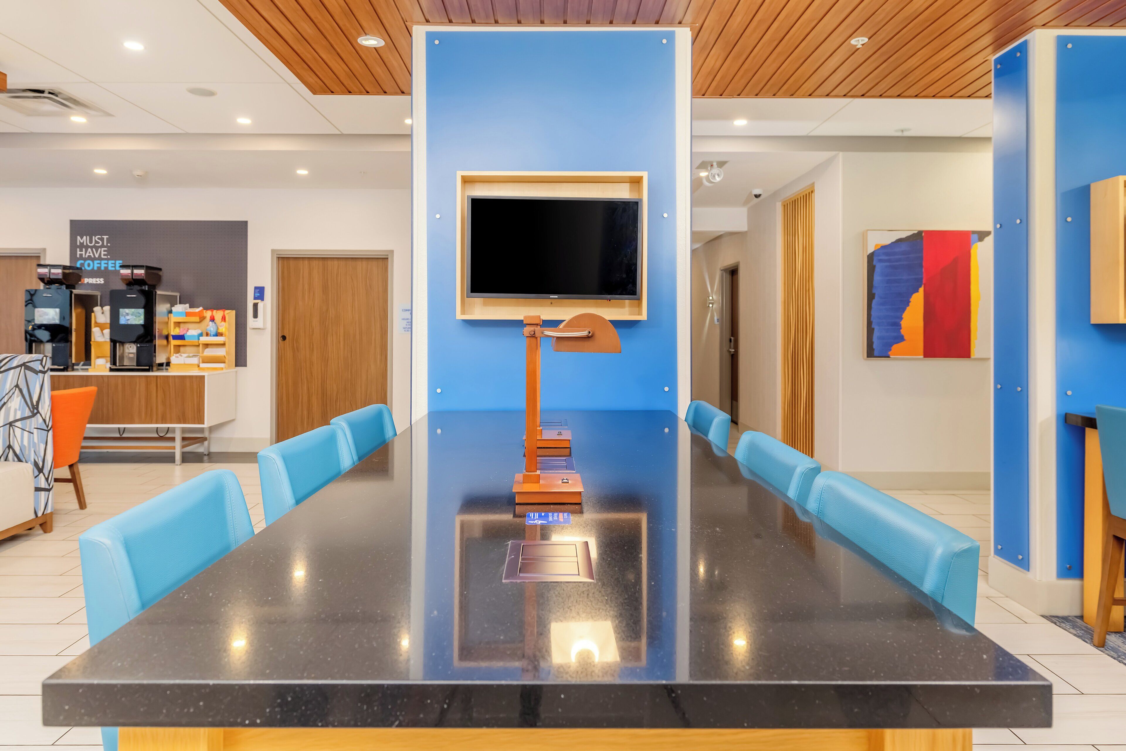 A modern conference room with a long table, blue chairs, a mounted TV, and a coffee station in the background.