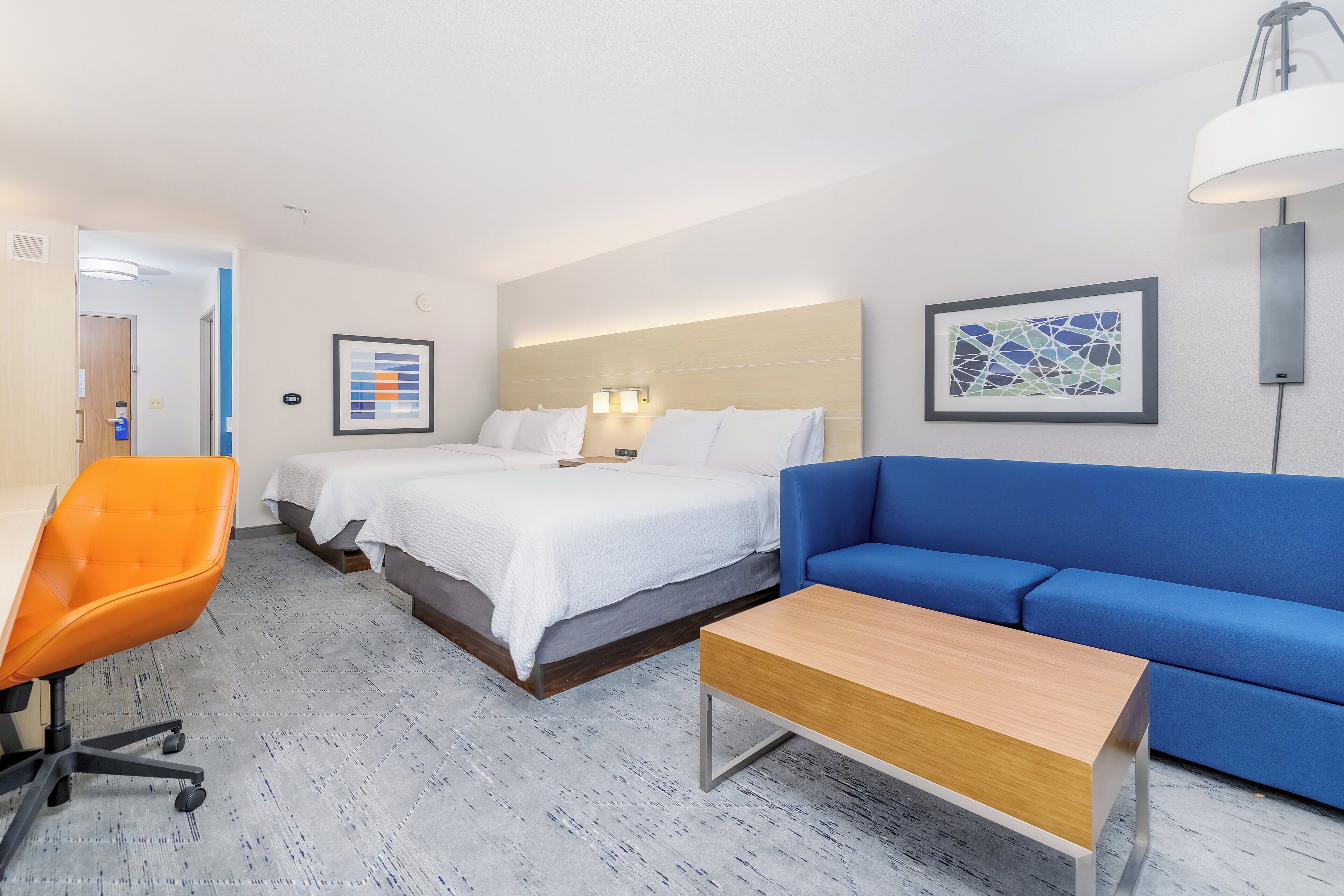 Quarto de hotel com cama, sofá azul, mesa de madeira, cadeira laranja e luminária fixada na parede.