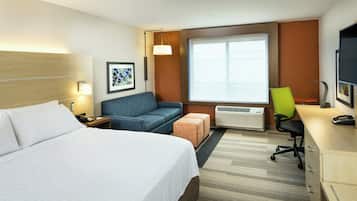 A hotel room with a large bed, a sofa, a desk with a chair, an air conditioning unit, and a window with blinds.