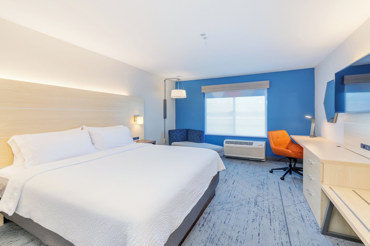 A hotel room with a bed, a desk, an orange chair, a window with blinds, and a wall-mounted TV.