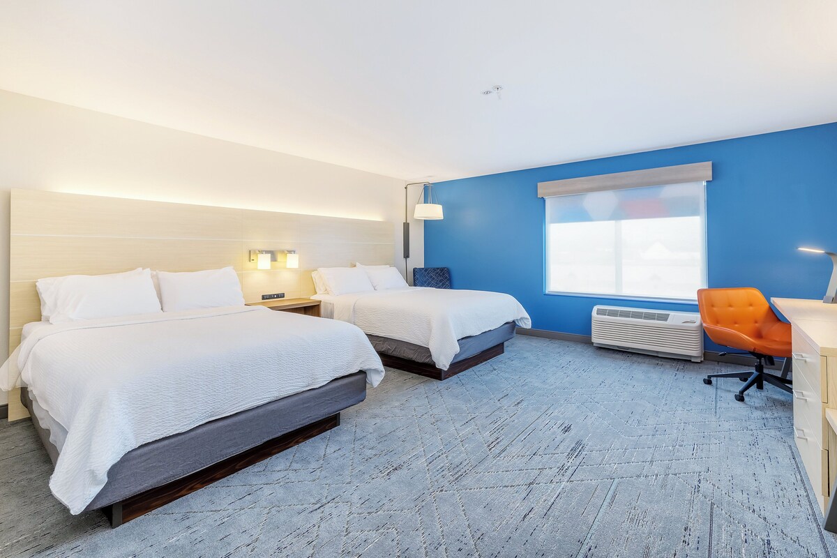 A hotel room with two beds, a desk, an orange chair, a window, and a wall-mounted air conditioner.