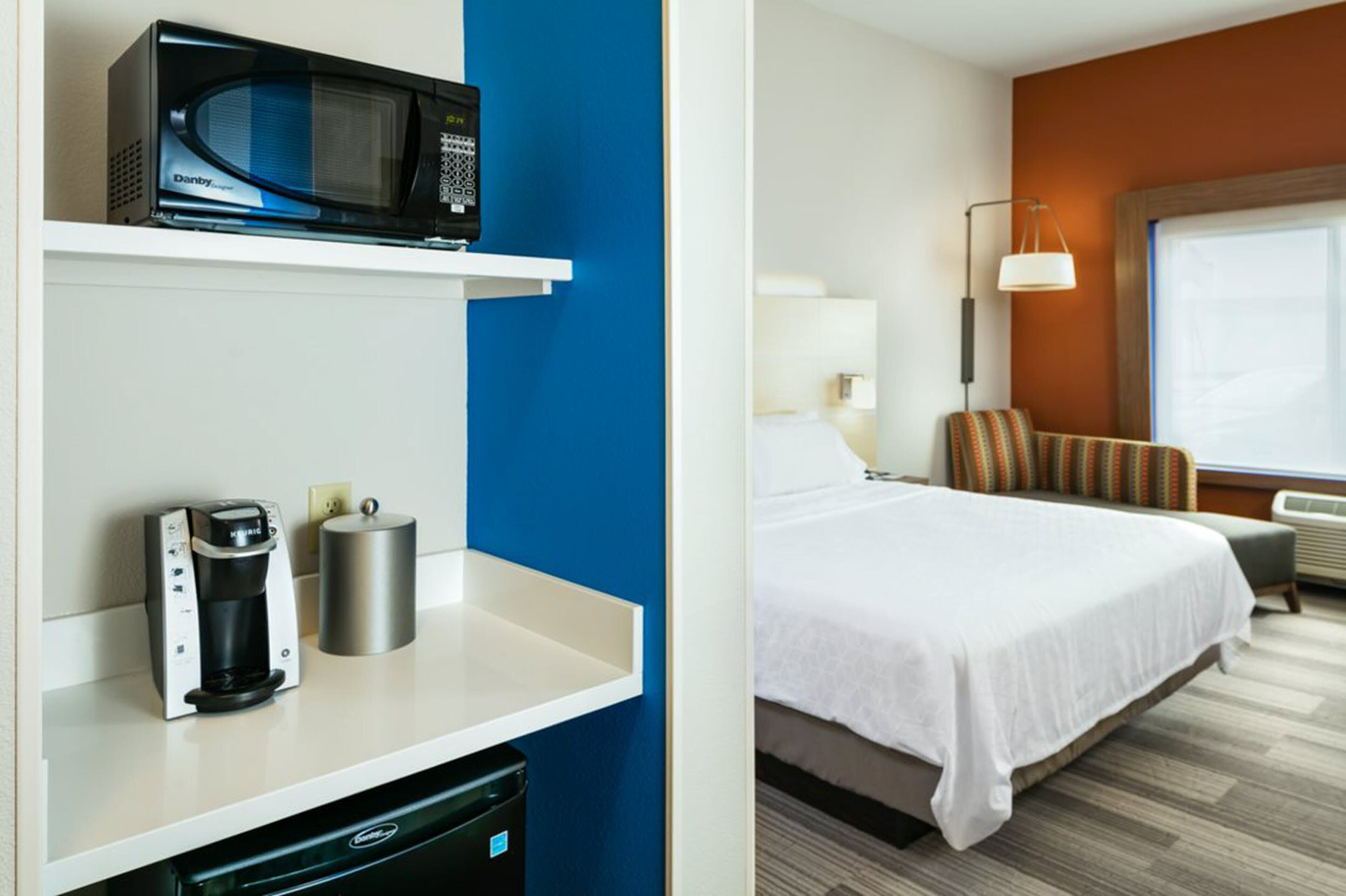 Habitación de hotel con cama, una silla, una lámpara y una kitchenette equipada con microondas, cafetera y cubo de basura.