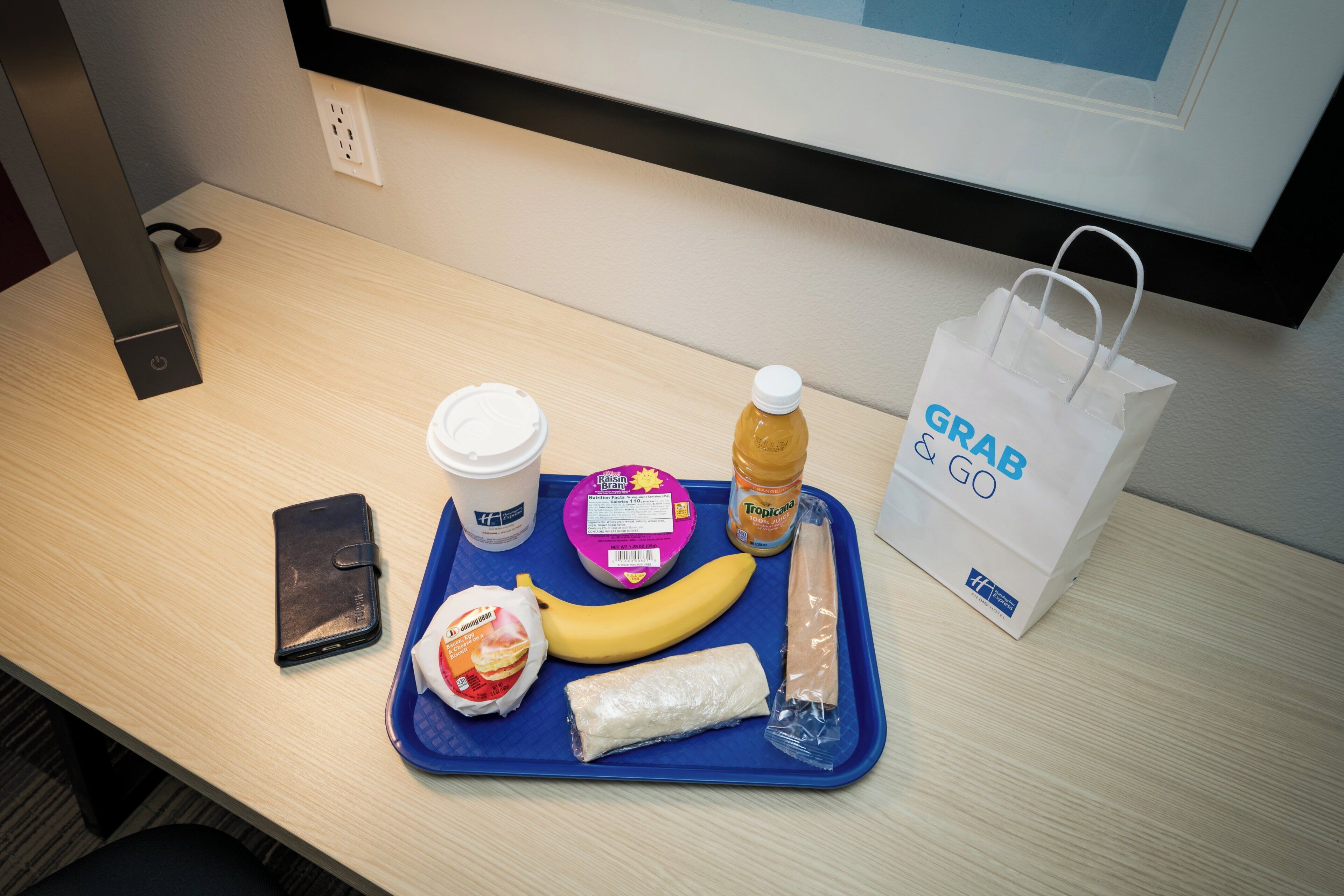 A tray with a banana, a bottle of juice, a wrapped item, a container of yogurt, and a cup, alongside a smartphone and a white bag with 'GRAB & GO' text.