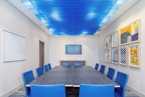 A conference room with a long table, blue chairs, a whiteboard, and artwork on the walls.