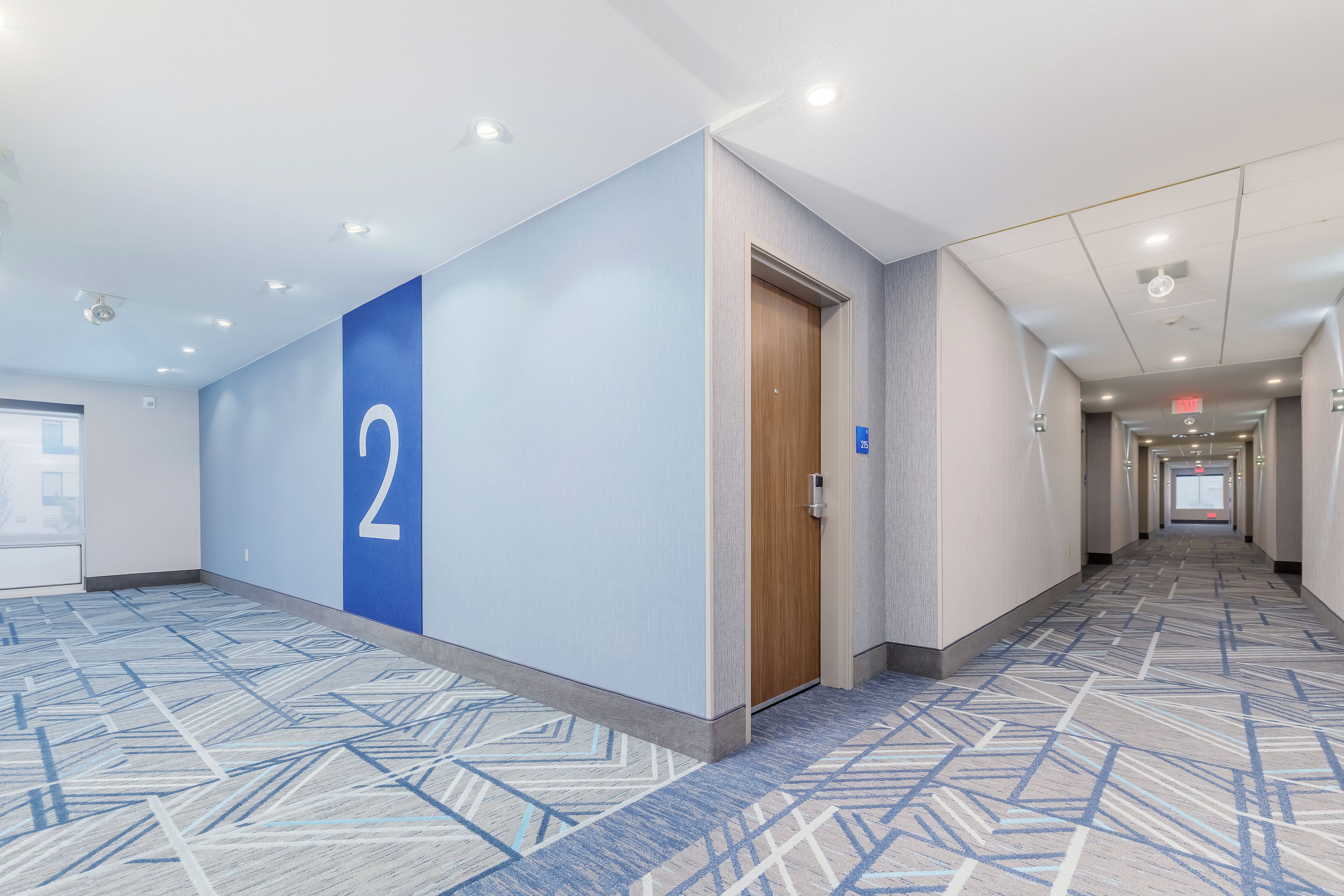 A hotel hallway with a blue wall featuring a large number '2', a wooden door, and patterned carpet.