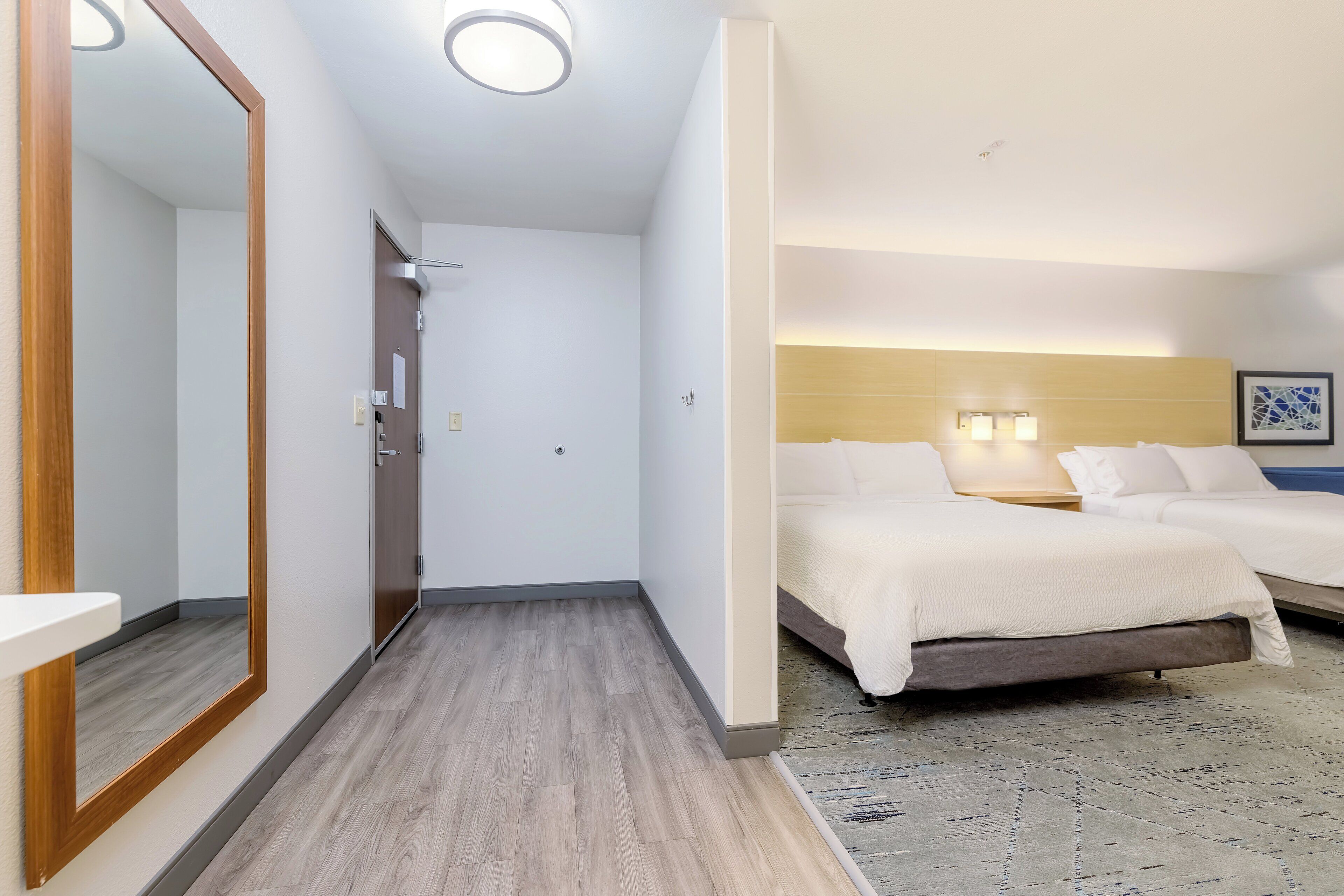 A hotel room with a wooden floor, a large mirror, a bed with white bedding, and a headboard with wall-mounted lights.