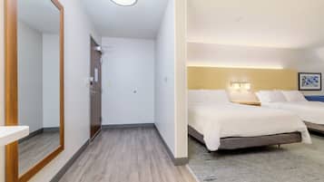 A hotel room with a wooden floor, a large mirror, a bed with white bedding, and a headboard with wall-mounted lights.