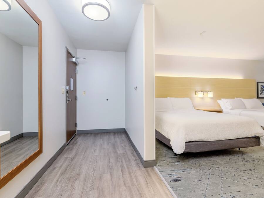 A hotel room with a wooden floor, a large mirror, a bed with white bedding, and a headboard with wall-mounted lights.
