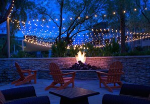 An outdoor seating area with Adirondack chairs and a fire pit, surrounded by a stone wall and string lights.